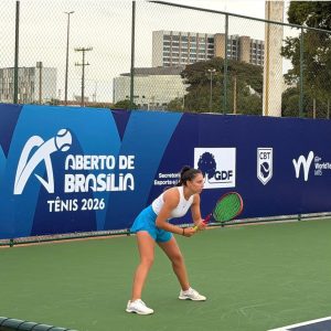 Tenista feminina em posição de expectativa para receber o saque em uma quadra rápida verde, com banners do Aberto de Brasília de Tênis 2026 e CBT ao fundo.