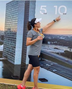 A corredora Juliana Sanches sorrindo, de boné e medalha no peito, apontando para a marca de 10 km em um painel com a foto do Congresso Nacional de Brasília no Eixão do Lazer.