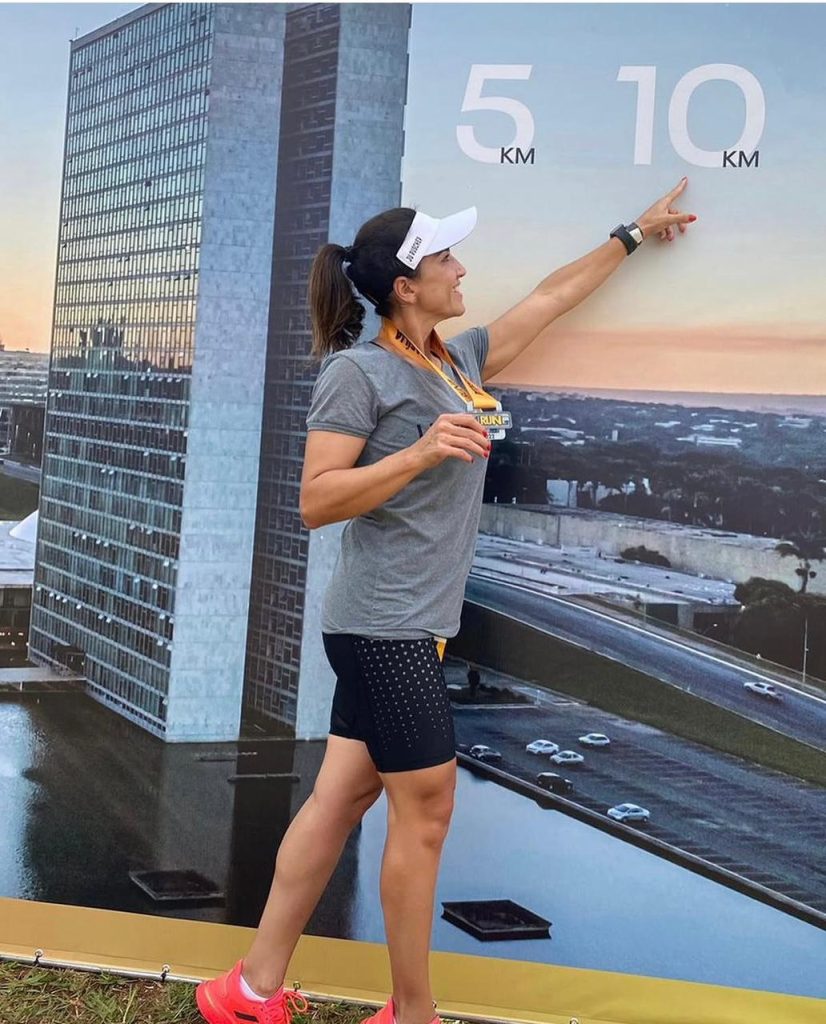 A corredora Juliana Sanches sorrindo, de boné e medalha no peito, apontando para a marca de 10 km em um painel com a foto do Congresso Nacional de Brasília no Eixão do Lazer.