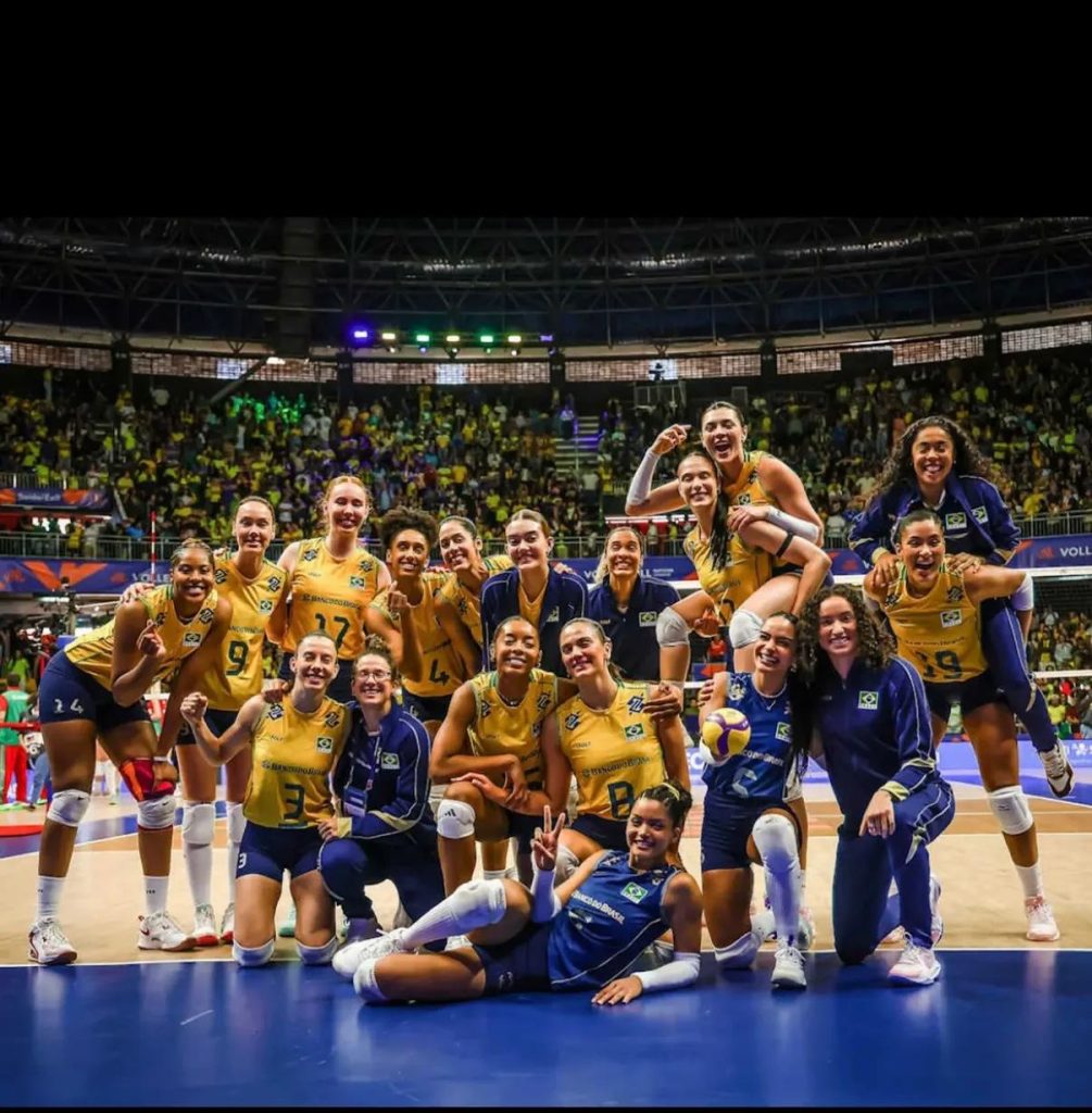 Elenco completo da Seleção Brasileira Feminina de Vôlei reunido e sorrindo na quadra do Ginásio Nilson Nelson, com a torcida ao fundo ocupando as arquibancadas em Brasília.