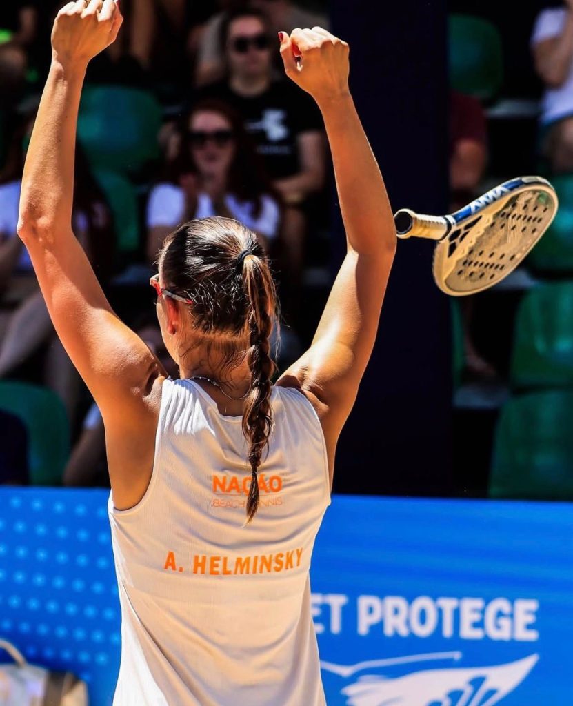 Atleta profissional Amanda Helminsky de costas, com os braços erguidos comemorando e a raquete jogada para o alto após vitória no Beach Tennis.