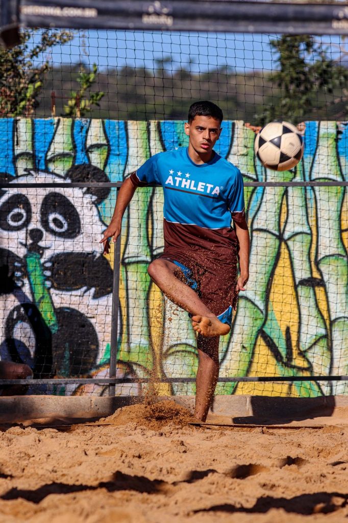 Atleta Guilherme Almeida dominando a bola de futevôlei com o pé na areia, em frente a um muro grafitado.
