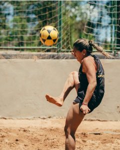 Atleta feminina saltando na areia para dominar uma bola amarela e preta de futevôlei durante torneio na Club.Co em Brasília.