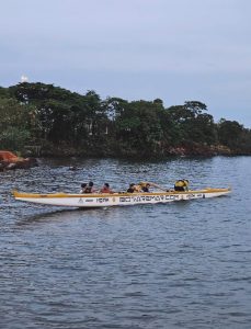 Uma canoa havaiana amarela e branca com várias pessoas remando no Lago Paranoá, em Brasília, durante o entardecer, com árvores ao fundo na margem oposta.
