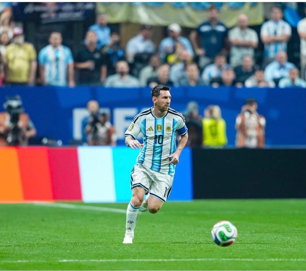 Lionel Messi, vestindo a clássica camisa 10 listrada em azul-celeste e branco da Argentina, é capturado em movimento correndo com a bola dominada nos pés durante uma partida. O fundo mostra as placas de publicidade do estádio e a torcida nas arquibancadas ligeiramente desfocadas.