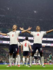 Em primeiro plano, os jogadores ingleses Harry Kane (camisa 9) e Jude Bellingham (camisa 10) de pé no gramado com os braços totalmente abertos e estendidos para os lados, olhando para o alto em um momento de celebração. Ao fundo desfocado, vê-se o companheiro Declan Rice (camisa 4) de costas e as arquibancadas cheias de torcedores.