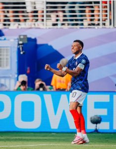 O atacante francês Kylian Mbappé, usando a camisa azul de número 10 e calção branco, é visto de perfil correndo e comemorando intensamente com os dois punhos cerrados e dentes cerrados.