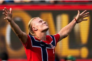 O centroavante norueguês Erling Haaland, em plano médio, aparece com os olhos fechados e a cabeça levemente inclinada para trás com uma expressão de êxtase e gratidão. Ele está com os braços totalmente erguidos para o alto e abertos, vestindo a camisa vermelha da seleção da Noruega. Ao fundo, um telão luminoso amarelo desfocado.