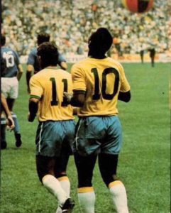 Foto histórica tirada de costas dos jogadores Pelé (número 10) e Rivelino (número 11), vestindo as camisas clássicas da Seleção Brasileira com calções azuis e meias brancas. Eles estão caminhando juntos no gramado de um estádio lotado.