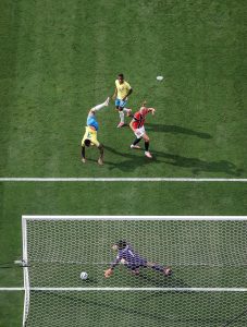 Foto tirada de cima mostrando uma jogada de futebol em um gramado verde. O atacante Erling Haaland, vestindo o uniforme vermelho da Noruega, observa a bola entrar no canto esquerdo do gol. O goleiro adversário, de uniforme roxo, está esticado no chão tentando a defesa, sem alcance. Próximo a eles, dois jogadores da Seleção Brasileira (um caído de ponta-cabeça após tentar o desarme e outro de pé observando) acompanham o lance.