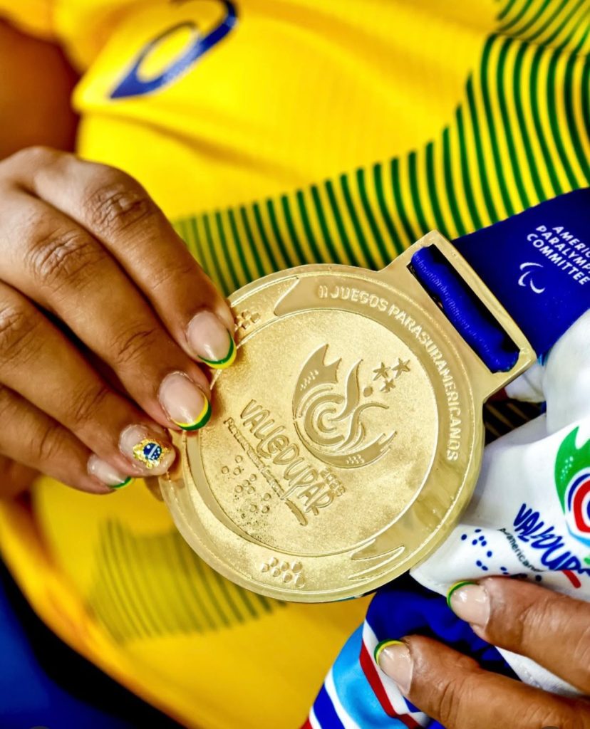 Foto em primeiro plano mostrando as mãos de uma atleta com unhas pintadas com detalhes em verde, amarelo e uma pequena bandeira do Brasil. Ela segura com orgulho uma grande medalha de ouro dourada com a inscrição 