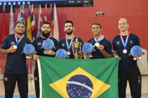 Cinco jogadores da seleção brasileira masculina de goalball posam alinhados lado a lado no pódio de uma competição esportiva. Eles vestem camisas azul-escuras oficiais da CBDV, usam medalhas no peito, seguram pequenos mascotes de pelúcia azuis e seguram juntos, esticada à frente deles, uma grande bandeira do Brasil.