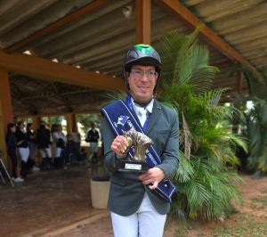 O paratleta de hipismo Sérgio Oliva sorri de pé, vestindo traje completo de hipismo cinza com calça branca e capacete de proteção preto com detalhes verdes. Ele segura um troféu em formato de cabeça de cavalo dourado e ostenta uma faixa azul de campeão cruzada no peito, no pátio da Sociedade Hípica de Brasília (SHBr).