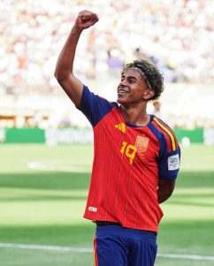 O jovem jogador espanhol Lamine Yamal sorri e comemora erguendo o punho direito cerrado em um estádio lotado. Ele usa a camisa vermelha número 19 da seleção da Espanha, tem aparelho nos dentes e uma faixa na cabeça prendendo o cabelo crespo com reflexos loiros. Crédito: FIFA / Reprodução Instagram