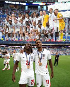 Montagem com duas imagens da seleção da Inglaterra comemorando a medalha de bronze na Copa do Mundo de 2026. Na parte superior, todo o elenco posa sorrindo em um pódio elevado. Na parte inferior, dois jogadores em primeiro plano posam orgulhosos no gramado exibindo suas medalhas de bronze no peito.