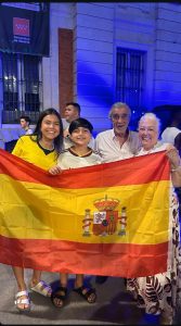Quatro pessoas da família Sanchez sorriem e posam juntas à noite na Praça do Sol, em Madrid, segurando bem aberta à frente delas uma grande bandeira oficial da Espanha. Entre elas estão duas crianças e dois idosos celebrando o título mundial.