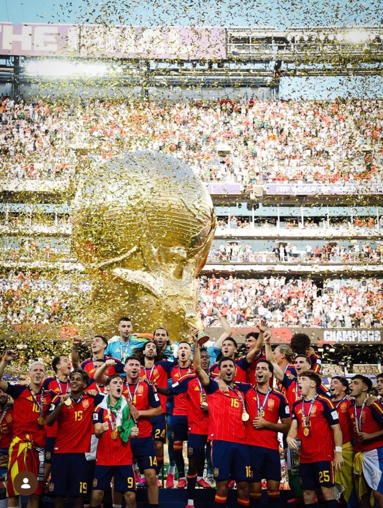 Jogadores da seleção espanhola de futebol masculino gritam e celebram em um pódio sob uma chuva intensa de confetes dourados. Ao fundo, destaca-se uma enorme estrutura inflável dourada no formato do troféu da Copa do Mundo da FIFA diante de uma arquibancada lotada.