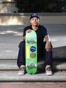 O skatista profissional Felipe Gustavo posa sentado em uma escadaria de concreto. Ele veste camiseta preta, calça escura, boné azul da Red Bull e segura em pé à sua frente um skate verde com o nome