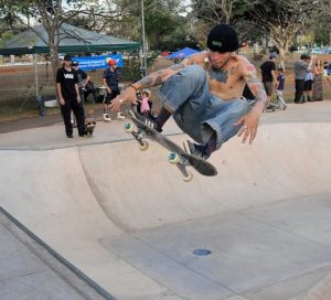 Skatista sem camisa, tatuado e usando touca preta e calça jeans larga, realiza uma manobra aérea alta sobre uma pista de concreto tipo bowl. Ele segura o skate no ar com a mão enquanto pessoas assistem ao fundo.