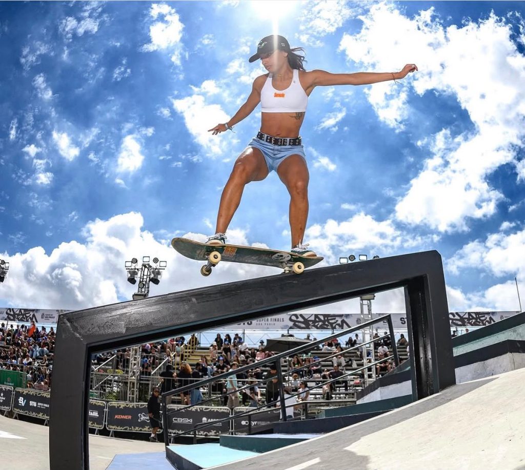 A skatista Kemily Sindy executa uma manobra equilibrando-se sobre um corrimão reto de metal preto em uma pista de skate street. Ela veste top branco, short jeans claro e boné, com um céu azul e nuvens brancas ao fundo.