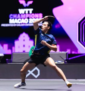 O mesatenista brasileiro Hugo Calderano com as pernas bem abertas e flexionadas em posição de combate, armando o braço direito com a raquete para rebater a bolinha durante uma partida. Ele veste uniforme azul com detalhes amarelos e calção preto.