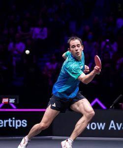 Hugo Calderano em movimento rápido pela quadra, olhando fixamente para a bolinha branca de tênis de mesa no ar enquanto prepara o golpe de forehand com a raquete. Ele veste camisa azul-turquesa e shorts preto.