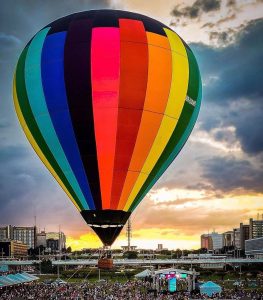 Um grande balão de ar quente com faixas verticais em cores do arco-íris sobe ao céu durante o pôr do sol. Abaixo do balão, vê-se a multidão reunida no gramado e a paisagem urbana com os prédios de Brasília.