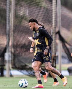 O jogador de futebol Neymar conduz uma bola branca e azul durante um treino no gramado. Ele veste uniforme preto do Santos com detalhes dourados, chuteiras chamativas e tem pernas bastante tatuadas.