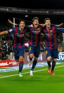 Os jogadores de futebol Luis Suárez, Neymar e Lionel Messi correm juntos com os braços abertos comemorando um gol, todos vestindo o uniforme azul e grená do Barcelona.