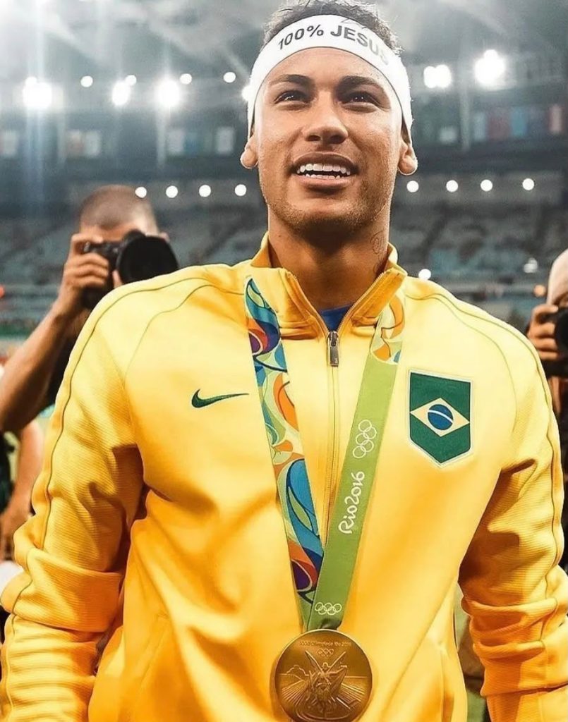 Neymar sorri usando a jaqueta amarela da delegação do Brasil, com a medalha de ouro dos Jogos Olímpicos Rio 2016 no peito e uma faixa branca na cabeça com a escrita 100% Jesus.