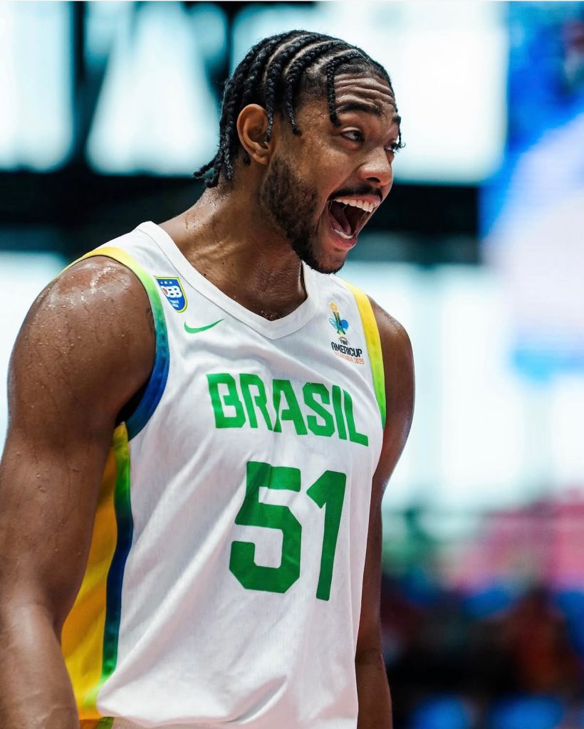 O jogador de basquete Bruno Caboclo vibrando intensamente com a boca aberta de vibração. Ele veste a regata branca da Seleção Brasileira com o número 51 verde no peito.
