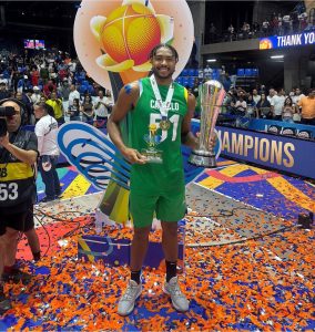 O pivô Bruno Caboclo sorrindo no pódio coberto de papéis picados coloridos. Ele segura um troféu prateado na mão esquerda, um prêmio individual na mão direita e usa medalha no peito vestindo uniforme verde.