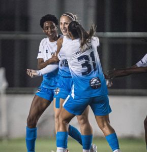 Três jogadoras de futebol do Real Brasília vestindo uniforme azul e branco abraçadas comemorando alegremente durante um jogo à noite.
