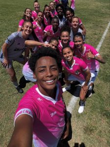 Foto em estilo selfie tirada por uma jogadora sorrindo no primeiro plano, acompanhada pelo grupo de atletas do Real Brasília reunidas no gramado sob o sol, todas vestindo uniforme rosa e branco.