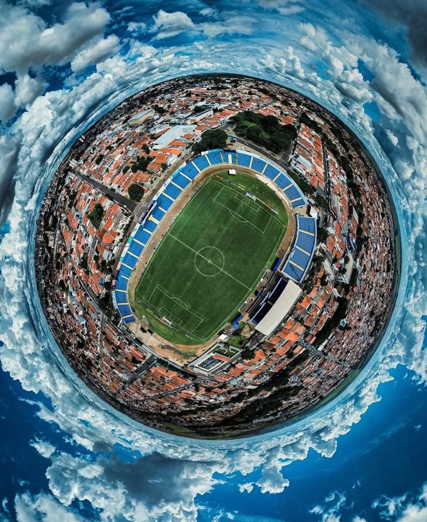 Fotografia panorâmica em formato esférico 360 graus mostrando um estádio de futebol com campo verde e arquibancadas azuis no centro, cercado pelo bairro e por nuvens no céu azul.