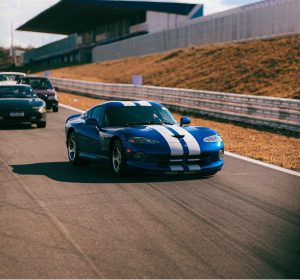 Carro esportivo Dodge Viper de cor azul brilhante com duas faixas brancas longitudinais acelerando na pista do autódromo, acompanhado por outros veículos ao fundo.