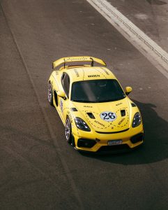 Vista superior de um carro esportivo Porsche amarelo de número 26, com aerofólio traseiro preto e detalhes estampados na lataria, rodando no asfalto do circuito.