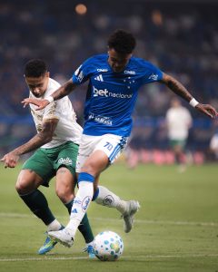 Dois jogadores disputando a bola no gramado: um vestindo a camisa azul e calção branco do Cruzeiro e o outro com camisa branca e calção verde.