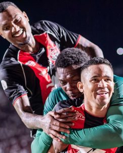Três jogadores do Vitória sorrindo e se abraçando entusiasmados durante a comemoração de um gol, vestindo camisas com as cores vermelha, preta e verde.