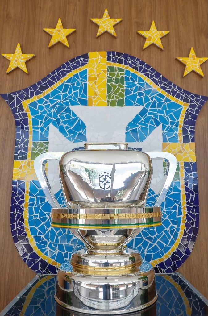 O troféu prateado da Copa do Brasil em primeiro plano sobre uma mesa espelhada, com o escudo mosaico da CBF em azul, amarelo e branco e estrelas douradas ao fundo.