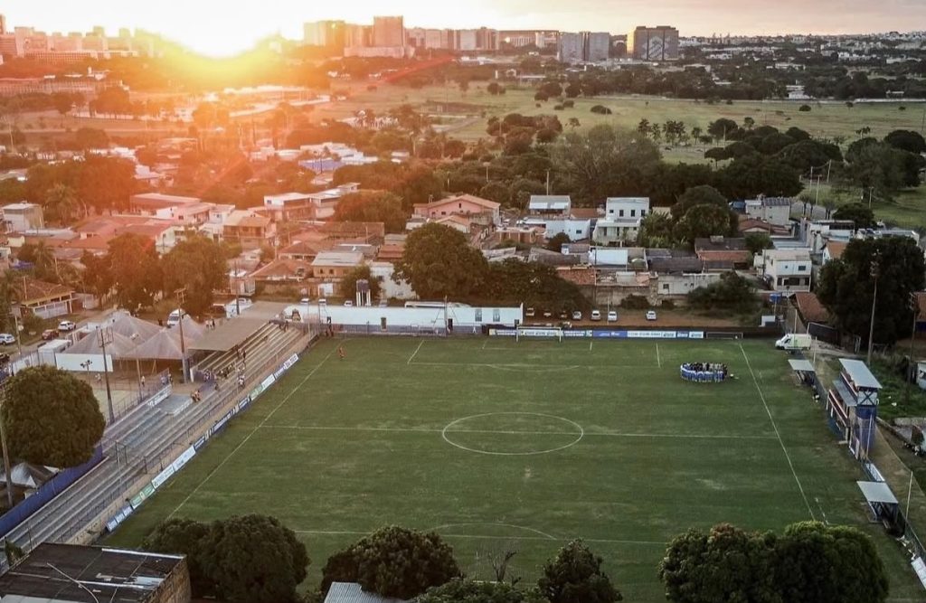 Vista panorâmica tirada por drone do Estádio Defelê, mostrando o gramado verde, as arquibancadas nas laterais e o bairro da Vila Planalto ao redor com o sol se pondo no horizonte.