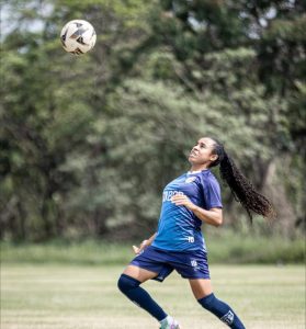 Jogadora de futebol com longo rabo de cavalo encaracolado olhando para o alto enquanto se prepara para dominar uma bola no ar. Ela veste o uniforme azul-escuro com o número 10 em destaque.