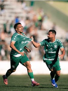 Dois jogadores de futebol do Gama comemorando um gol no gramado; o atleta à esquerda puxa a gola da própria camisa verde enquanto o companheiro ao lado corre gritando de emoção.