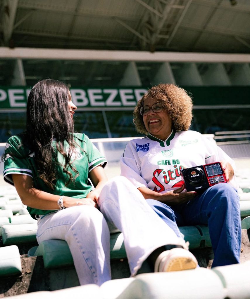 Duas torcedoras do Gama sorrindo e conversando na arquibancada do estádio; uma jovem com camisa verde e uma mulher com camisa retrô segurando um rádio portátil vermelho.