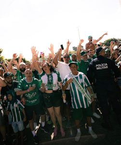 Grupo de torcedores do Gama reunidos na rua com braços levantados e rostos expressivos, vestindo camisas verdes e brancas do clube, com um policial militar ao lado.