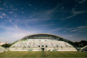 Visão ampla do Estádio Bezerrão a partir do gramado, mostrando a grande estrutura coberta da arquibancada sob um céu azul ensolarado com poucas nuvens.