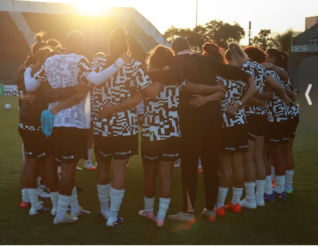 Jogadores e comissão técnica do Corinthians abraçados de costas em roda no gramado ao pôr do sol, vestindo camisa de treino estampada em preto e branco.