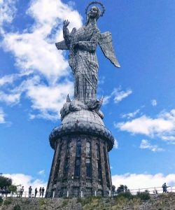 Fotografia em ângulo baixo mostrando a enorme estátua prateada e alada da Virgem Maria no topo do morro El Panecillo, em Quito. Ela está sobre um pedestal de pedra redondo. O céu está azul brilhante com nuvens brancas dispersas. Pequenas silhuetas de pessoas são visíveis na base, destacando a escala monumental da escultura.