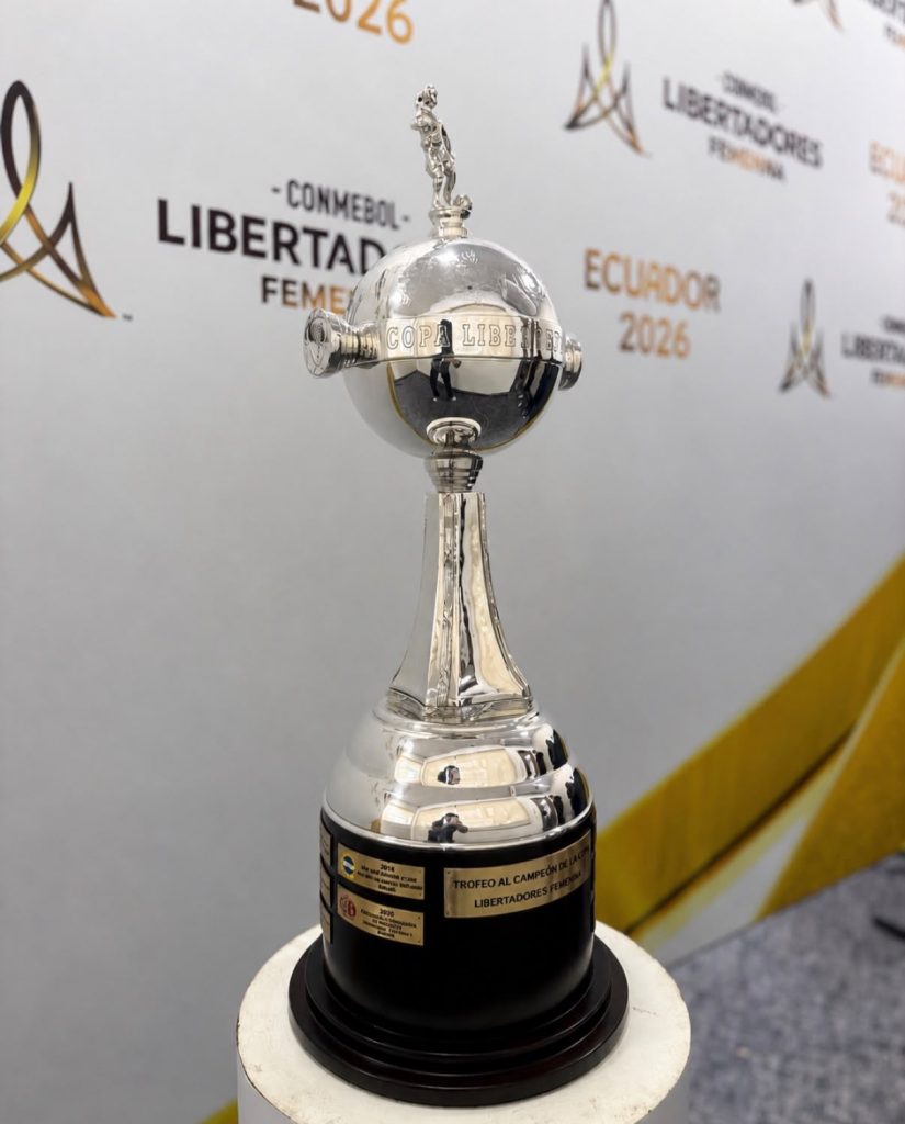 Uma foto em close-up do troféu da Copa Libertadores Feminina. A taça é prateada com uma base preta redonda contendo pequenas placas douradas de campeões anteriores. O corpo principal é uma esfera de metal polido encimada por uma pequena figura dourada de um jogador de futebol. O fundo é um banner de evento com a inscrição