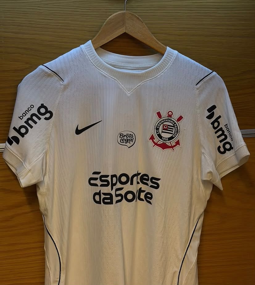 Camisa oficial branca do Corinthians pendurada num cabide de madeira, exibindo o logótipo da patrocinadora Esportes da Sorte impresso no centro do peito.
